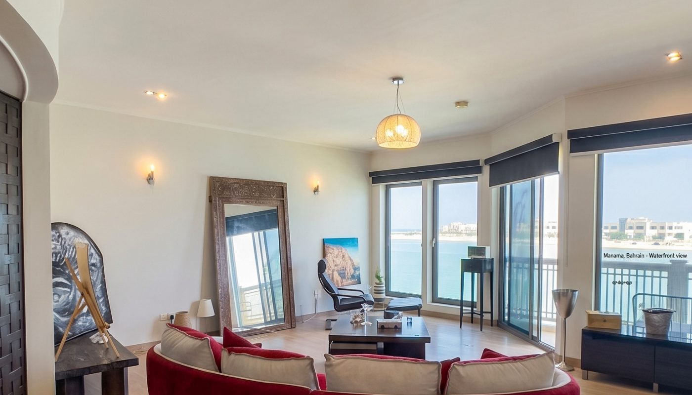 A modern living room with a curved red sofa, large windows with water views, light wood flooring, and contemporary decor including a large mirror and artwork.