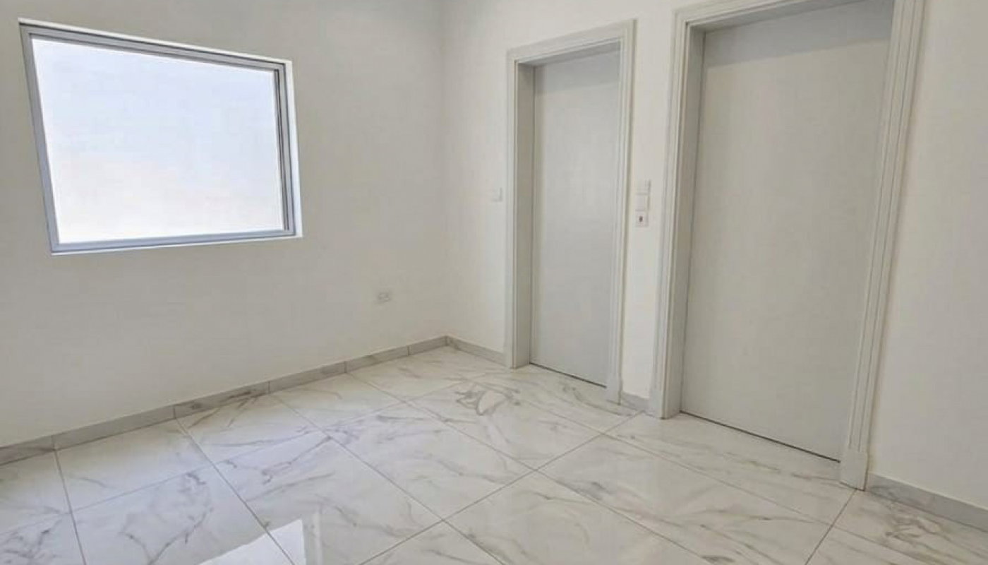 A bright, empty room with white marble tile flooring, a single window, and two closed doors on a white wall.
