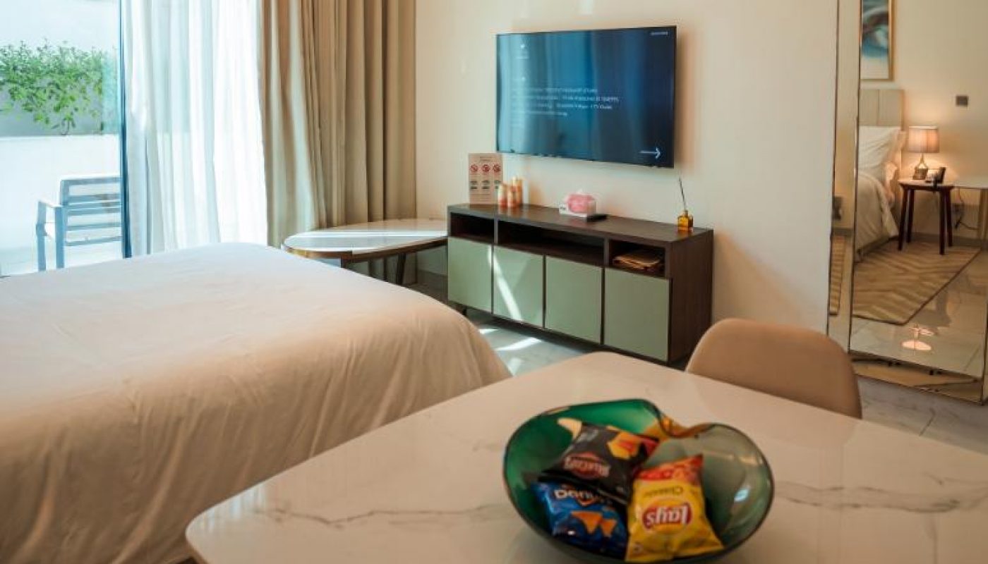Modern hotel room with a bed, TV, desk, marble table, bowl of snacks, pendant lights, large window with curtains, and a partial view of a balcony.
