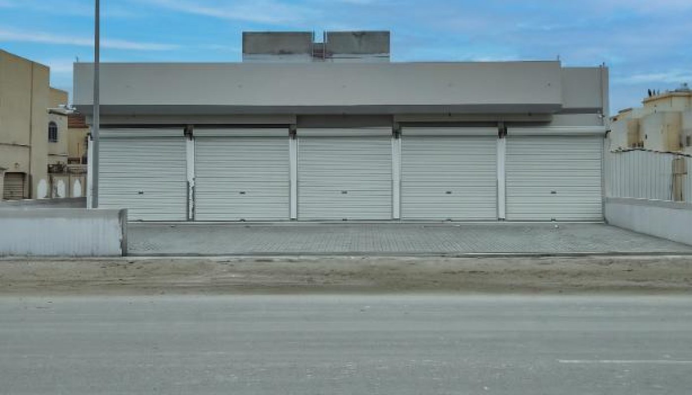 Gray commercial building with four closed white roll-up doors, empty parking area, and a street in the foreground under a blue sky.