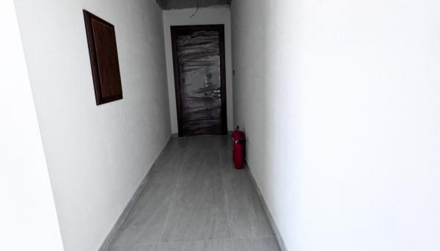 A narrow hallway with white walls and gray tiled floor leads to a closed wooden door; a fire extinguisher is placed on the floor near the door.