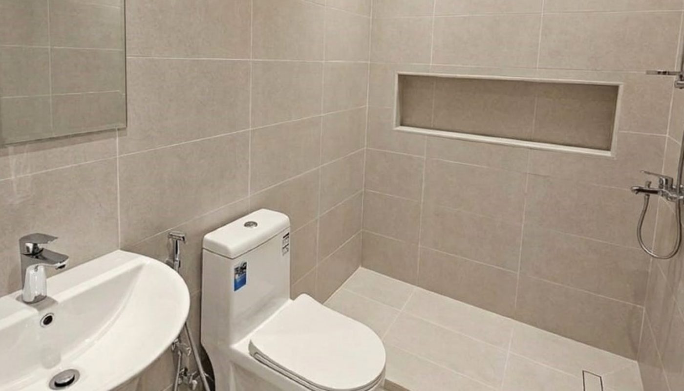 A modern bathroom with a wall-mounted sink, a toilet, a handheld bidet, a walk-in shower with a recessed shelf, and beige tiled walls and floor.