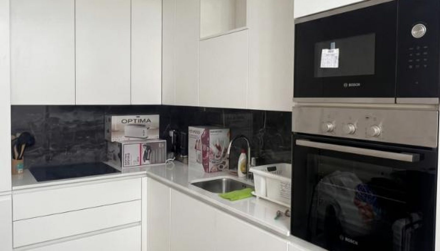 Modern white kitchen with built-in appliances, black backsplash, and light wood flooring. Box and kitchen items are on the countertop near the sink.