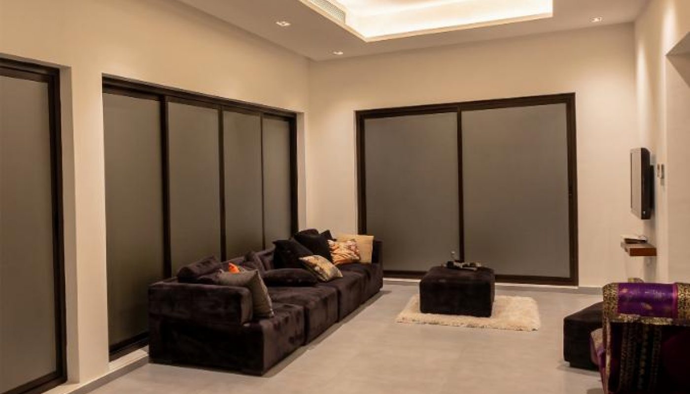 Modern living room with black sectional sofa, several cushions, a small ottoman, light gray tile flooring, recessed ceiling lights, and large windows with frosted glass.