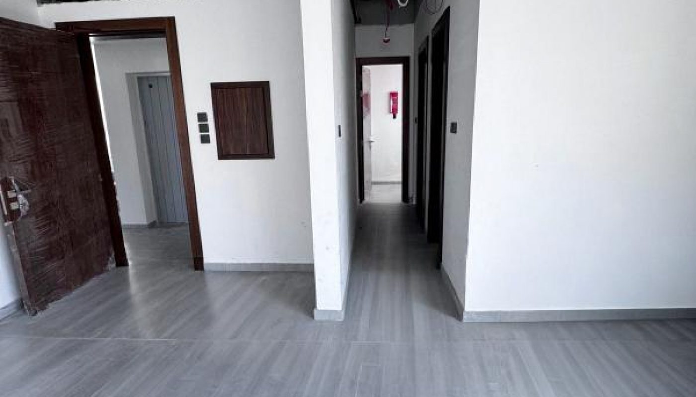 Interior of an unfinished apartment with exposed ceiling wiring, white walls, light gray flooring, and doorways leading to other rooms.