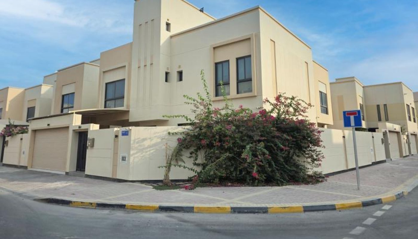 Modern beige two-story house on a corner lot with a garage, large windows, and flowering vines along the wall under a partly cloudy sky.