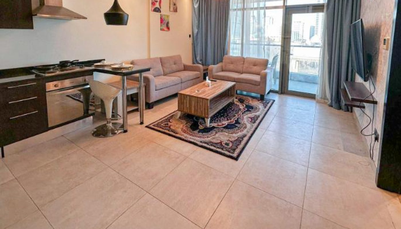 Modern open-plan living room with kitchen, beige tile flooring, two sofas, a coffee table, wall-mounted TV, and large windows with city view. Contemporary lighting and decor.