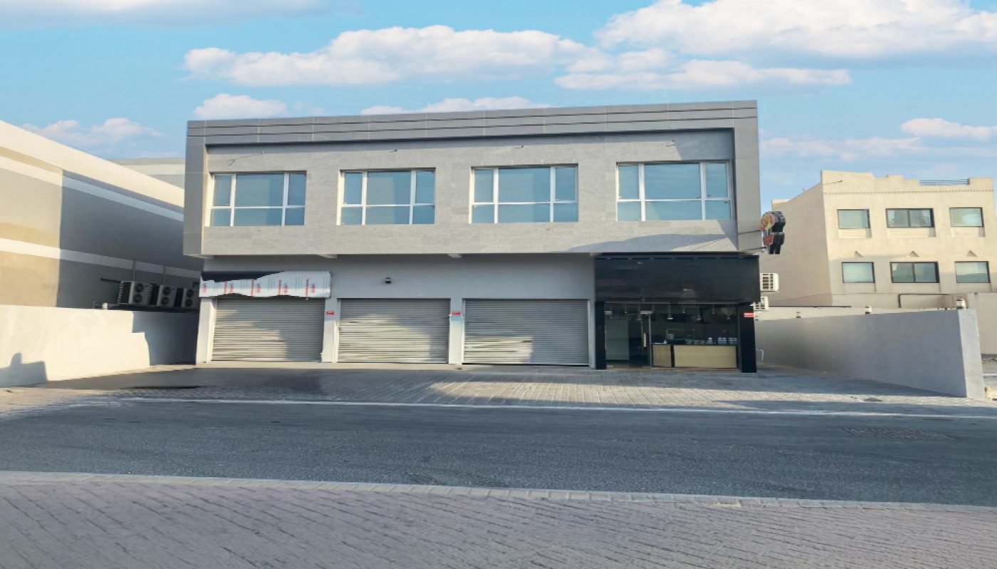 A modern two-story gray building with large windows and closed garage doors, situated on a paved street under a partly cloudy sky.
