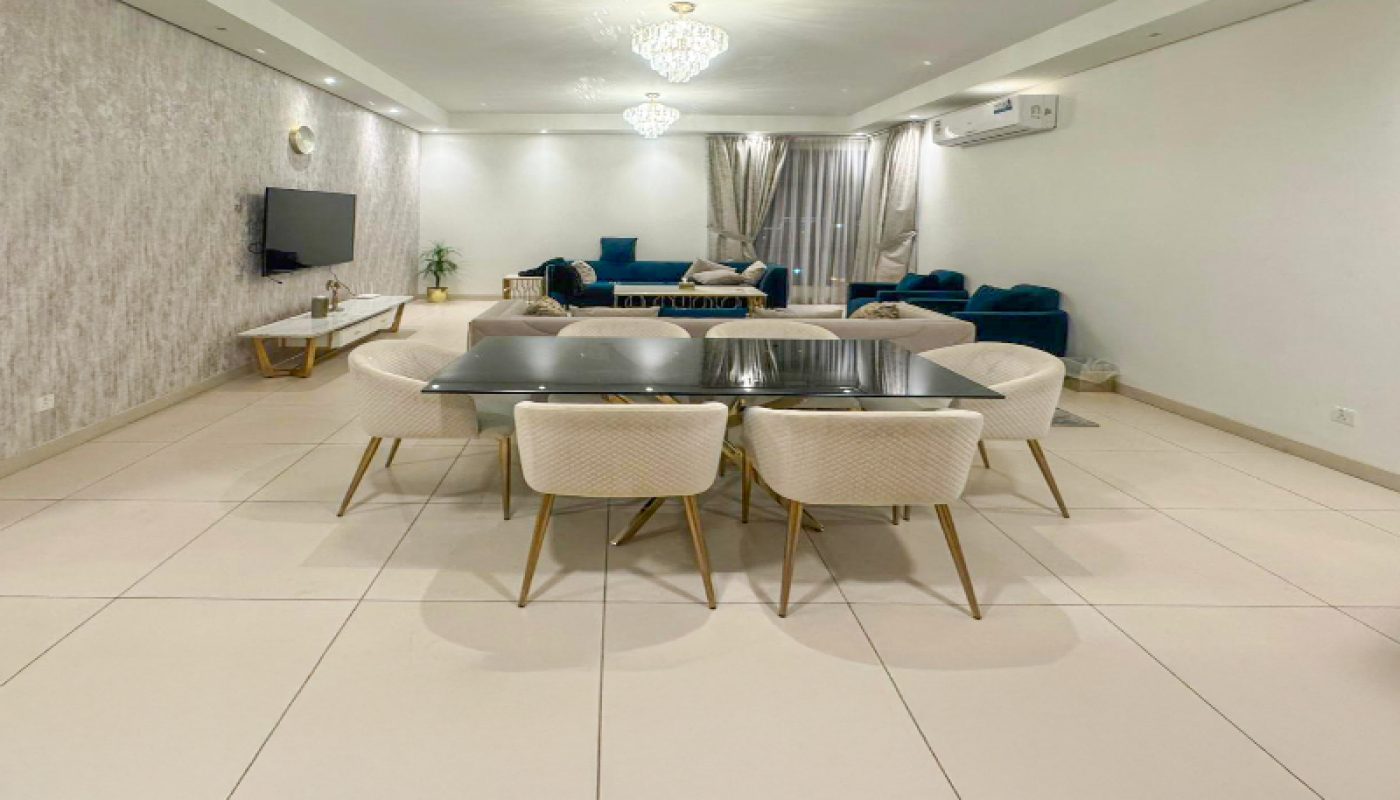 Modern open-plan living and dining area with a black dining table, six white chairs, a sofa set, wall-mounted TV, and chandelier lighting.