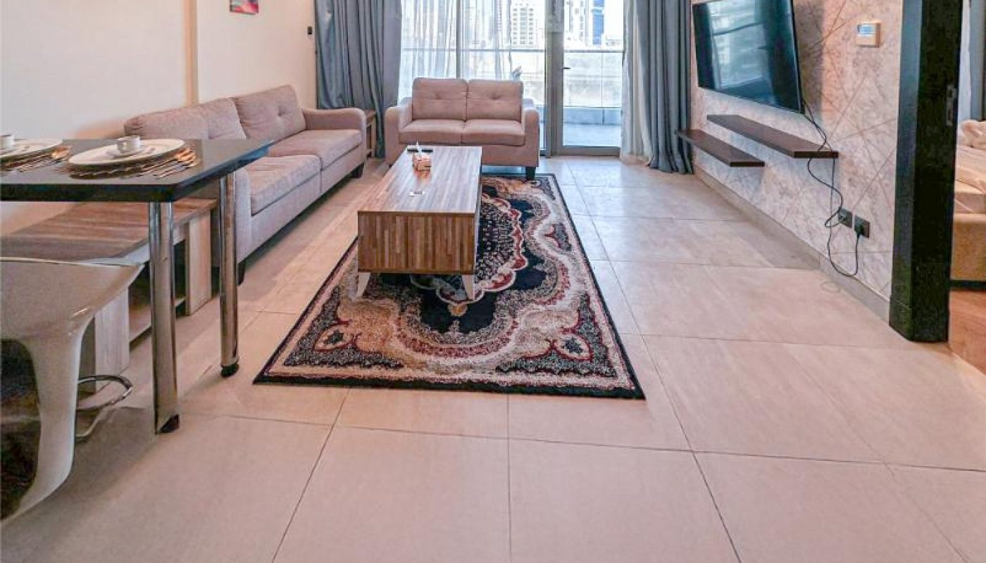 Modern living room with two sofas, a TV on a tiled wall, a coffee table on a patterned rug, and a dining area with a pendant light. Large windows with curtains let in natural light.