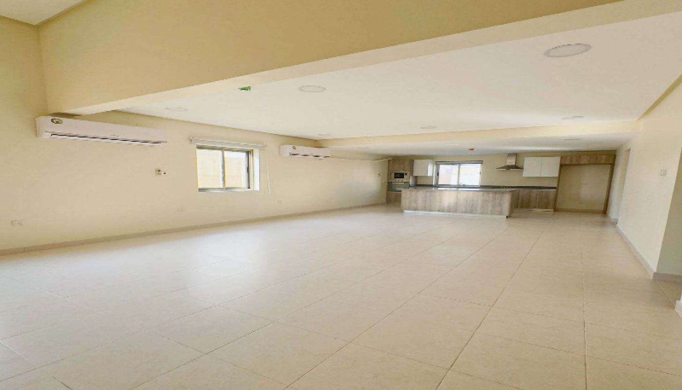 Spacious, empty room with tiled floors, beige walls, two windows, three air conditioners, and an open kitchen area in the background.
