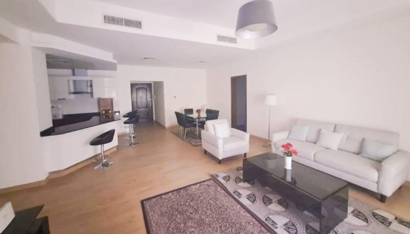 Modern open-plan living room with light wood flooring, white sofa set, black coffee table, dining area, kitchen with bar stools, and a decorative rug.