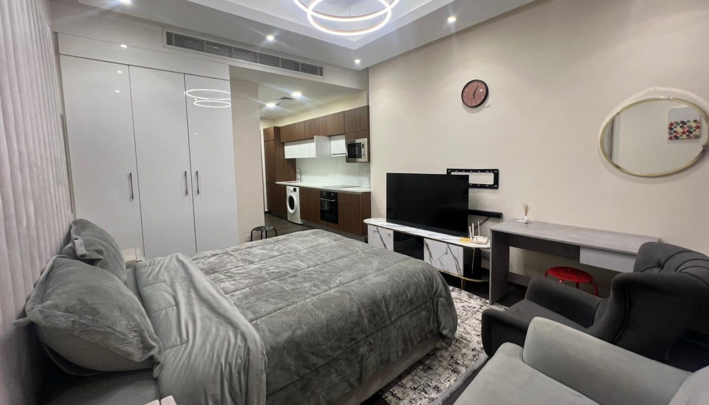 Modern studio apartment with a bed, armchair, TV, desk, wardrobe, and a kitchen area; white and gray decor with bright ceiling lights and minimal wall decorations.