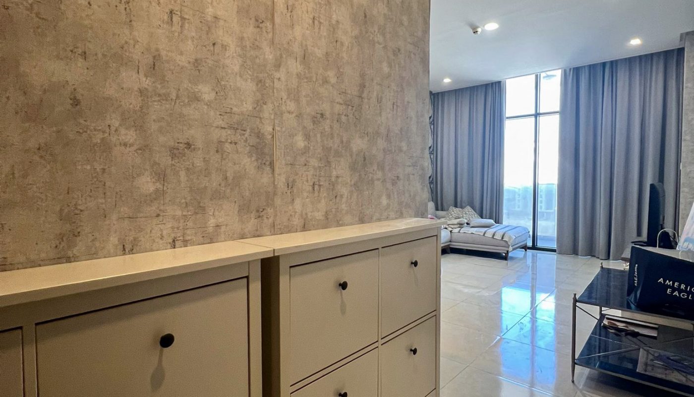 A hallway with a beige cabinet leads to a bright living room with large windows, tiled floors, and neutral-colored furniture.