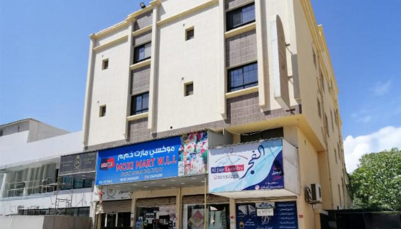 A four-story beige building with a grocery store and pharmacy on the ground floor, located on a quiet street under a clear sky.