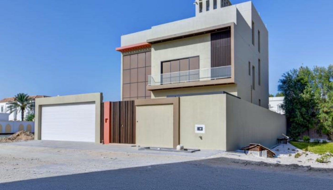 A modern three-story house with a flat roof, large windows, a two-car garage, and a driveway, set on a sunny day with clear skies.