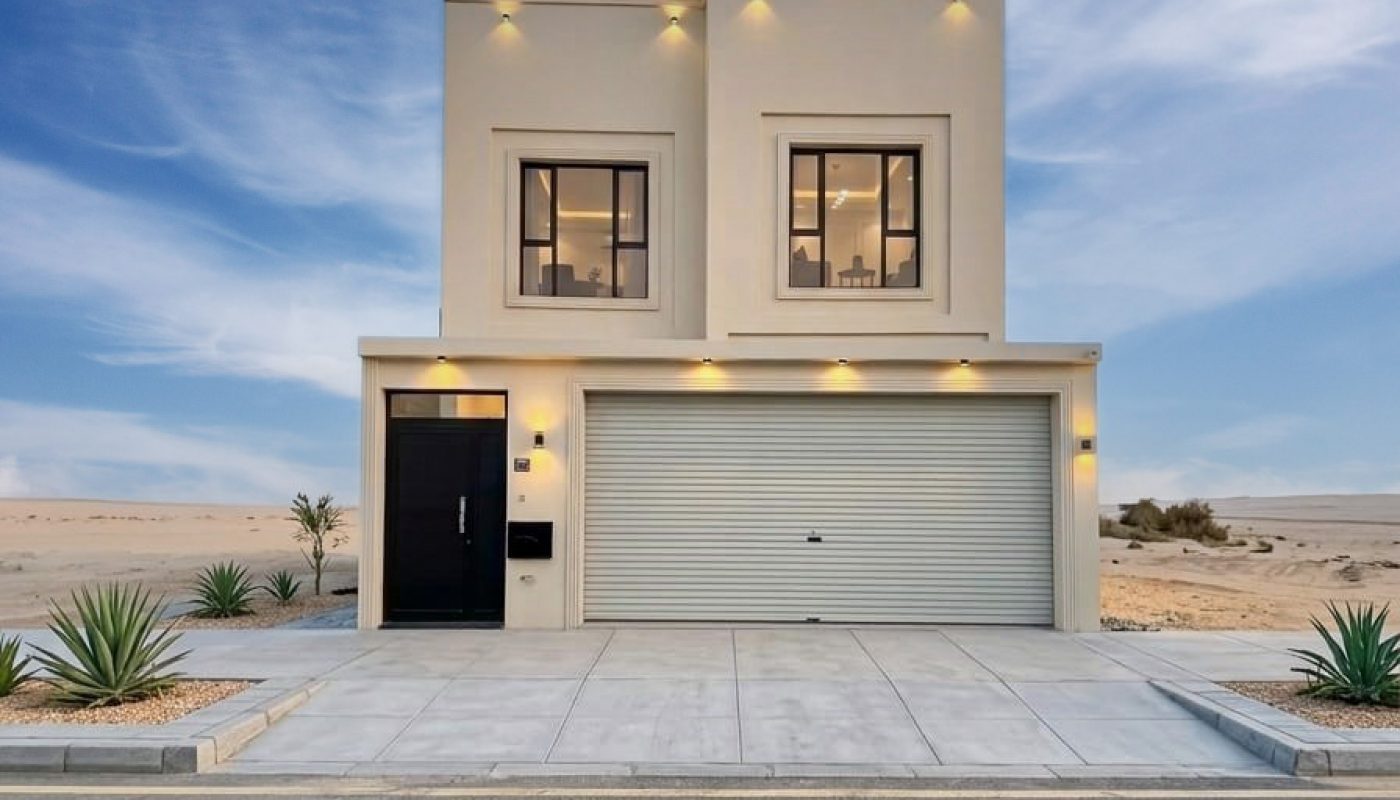 Modern two-story house with a flat roof, beige exterior, large windows, and a closed garage, set against a desert landscape under a partly cloudy sky.