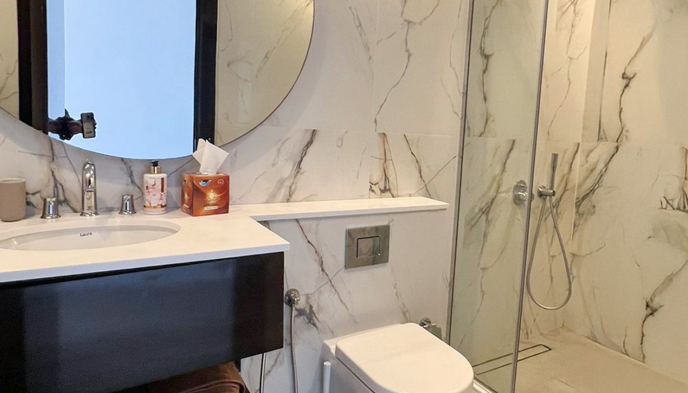 Modern bathroom with marble walls, a round mirror, sink with cabinet, wall-mounted toilet, glass shower enclosure, and a grey floor mat.