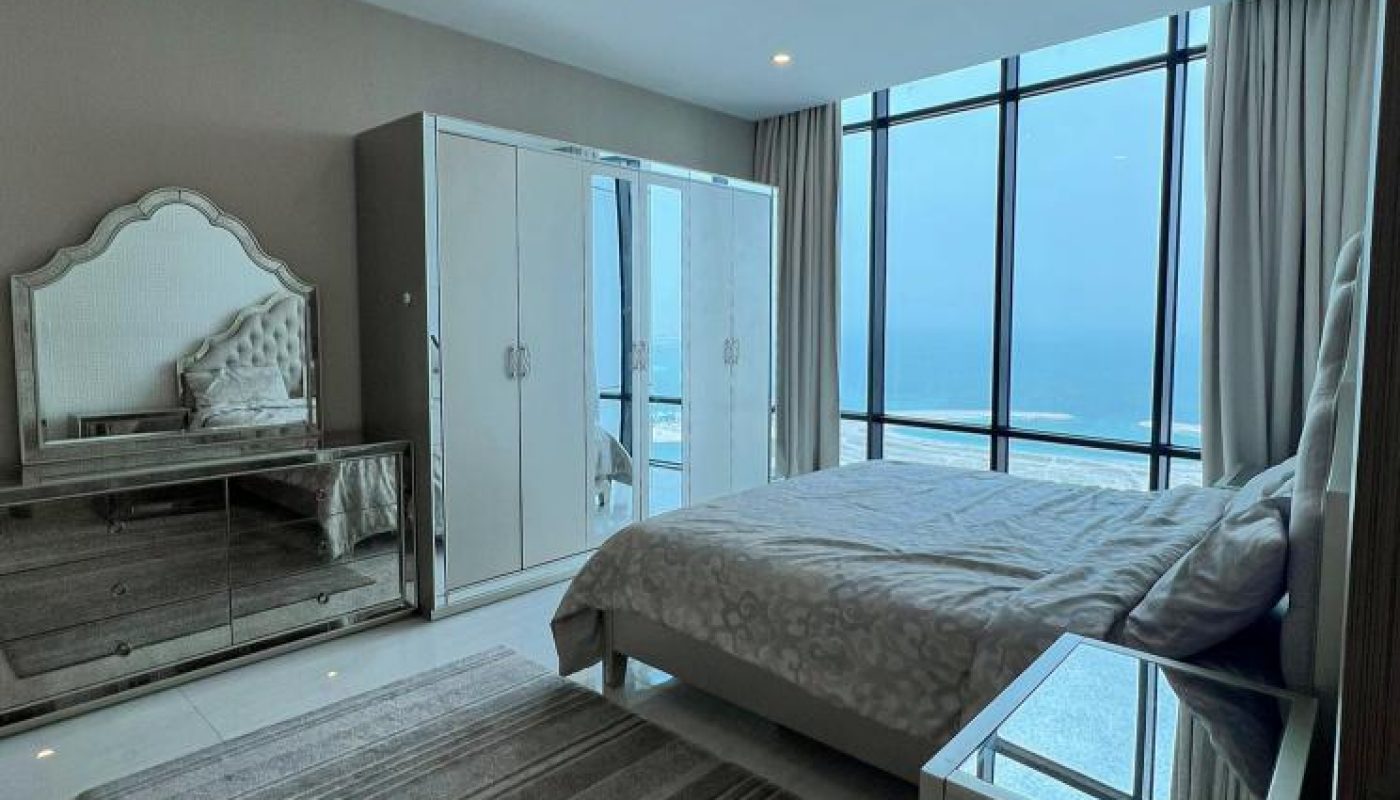 Modern bedroom with a large window overlooking the sea, featuring a bed, mirrored wardrobe, dresser with mirror, and a striped rug on a marble floor.