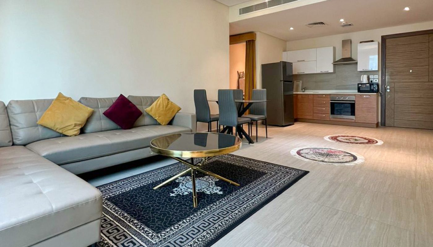 Modern open-plan living room and kitchen with a sectional sofa, dining table, coffee table, and built-in appliances. Decor includes cushions, rugs, and recessed ceiling lighting.