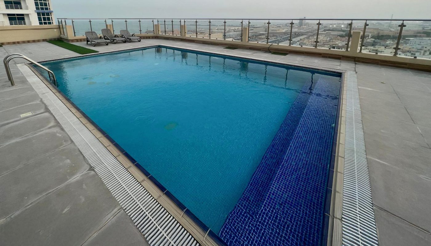 Rectangular rooftop swimming pool with metal railings and cityscape view in the background; several lounge chairs are arranged along one side.