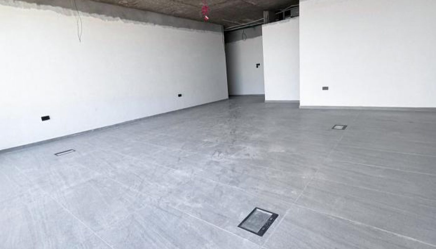 Empty room with unfinished concrete ceiling, white walls, grey tiled floor, and visible electrical outlets. Ceiling wiring and ducts are exposed; no furniture is present.
