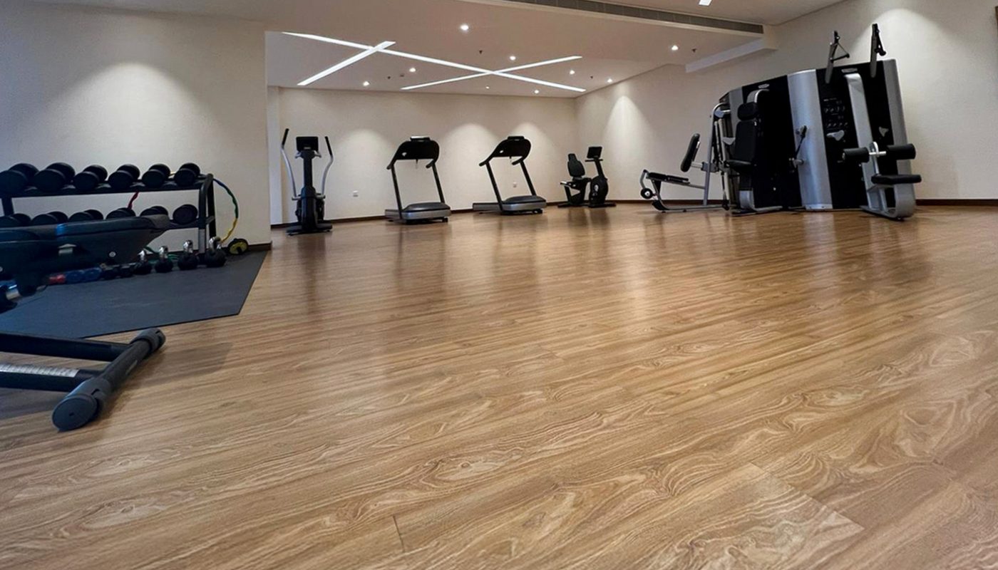 Spacious gym with wooden flooring, treadmills, stationary bikes, weightlifting benches, dumbbells, and a multi-station workout machine against white walls.