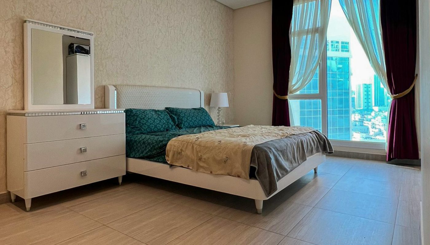 Modern bedroom with a bed, two bedside tables, a dresser with a mirror, large floor tiles, and a window with city views and curtains.