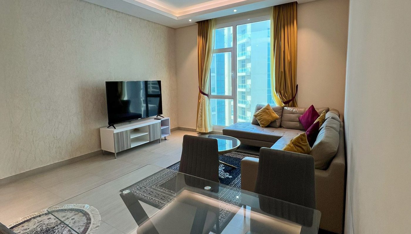 Modern living room with a glass dining table, gray chairs, L-shaped sofa, TV on a stand, large window with curtains, and city view.