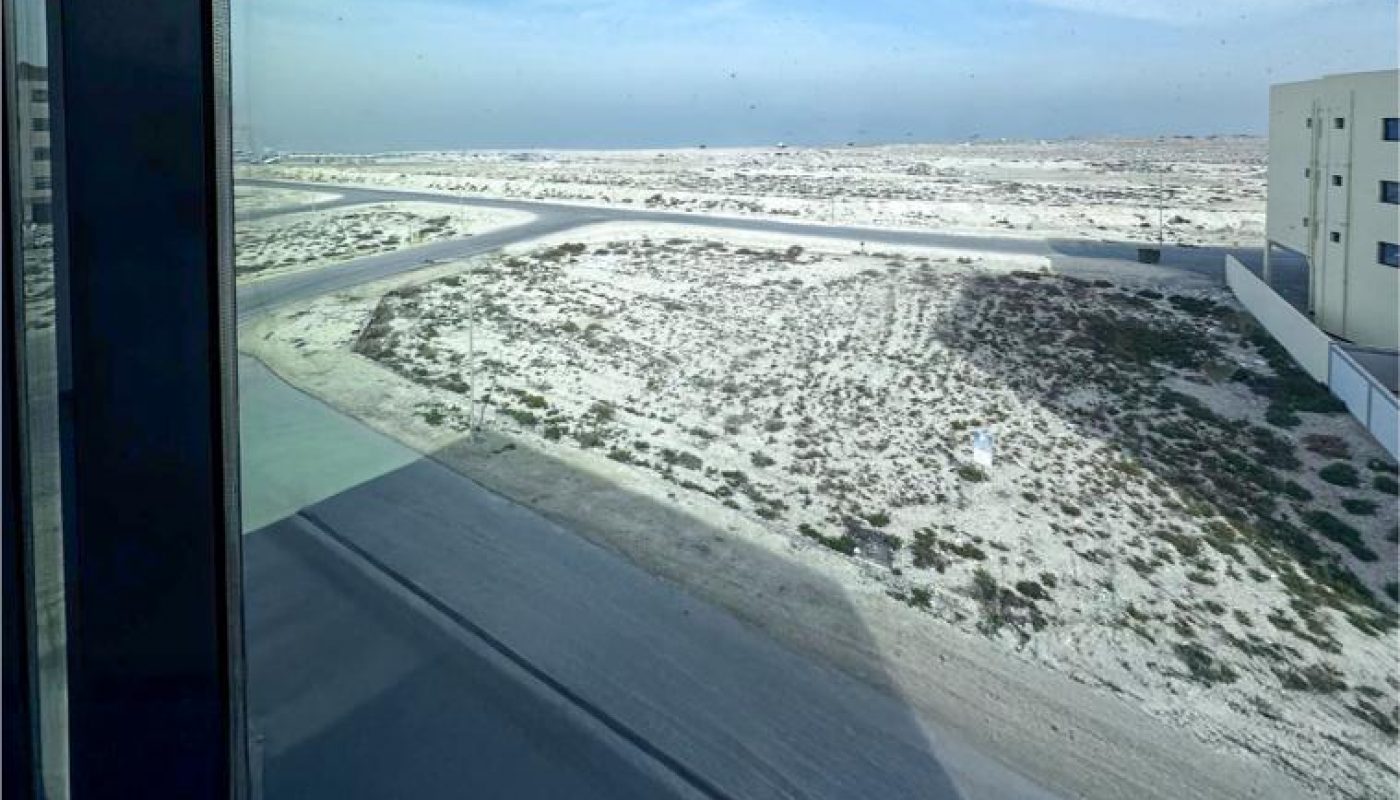 View from a window showing a paved road curving through a sandy, sparsely vegetated landscape with some buildings on the side under a blue sky.