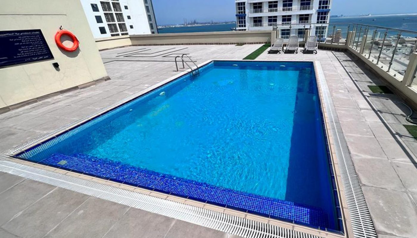 Rectangular outdoor swimming pool on a rooftop deck, surrounded by tall buildings, with clear blue sky and ocean visible in the background.