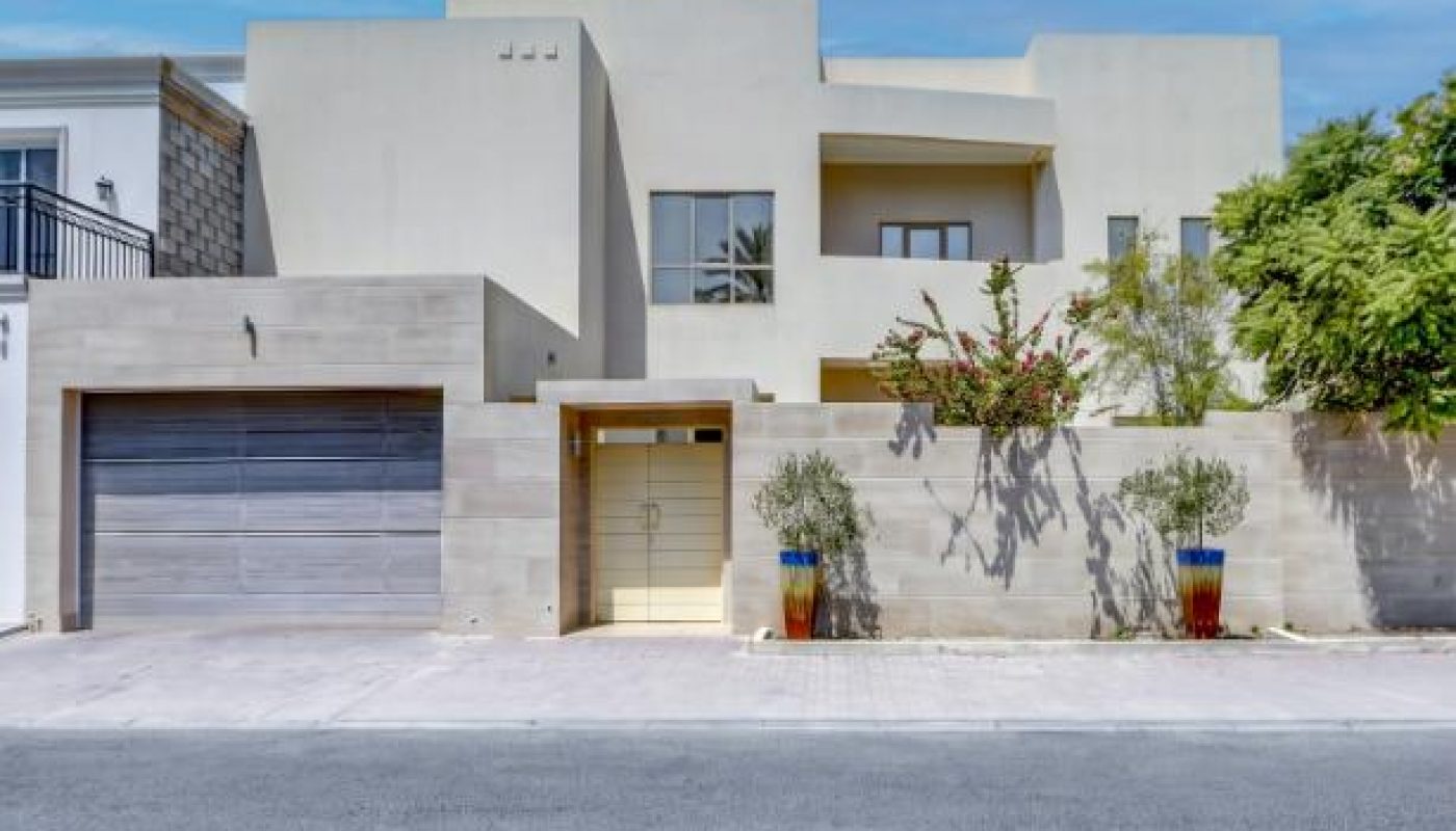 Modern two-story house with a flat roof, beige exterior, large garage door, and potted plants flanking the entrance on a sunny day.