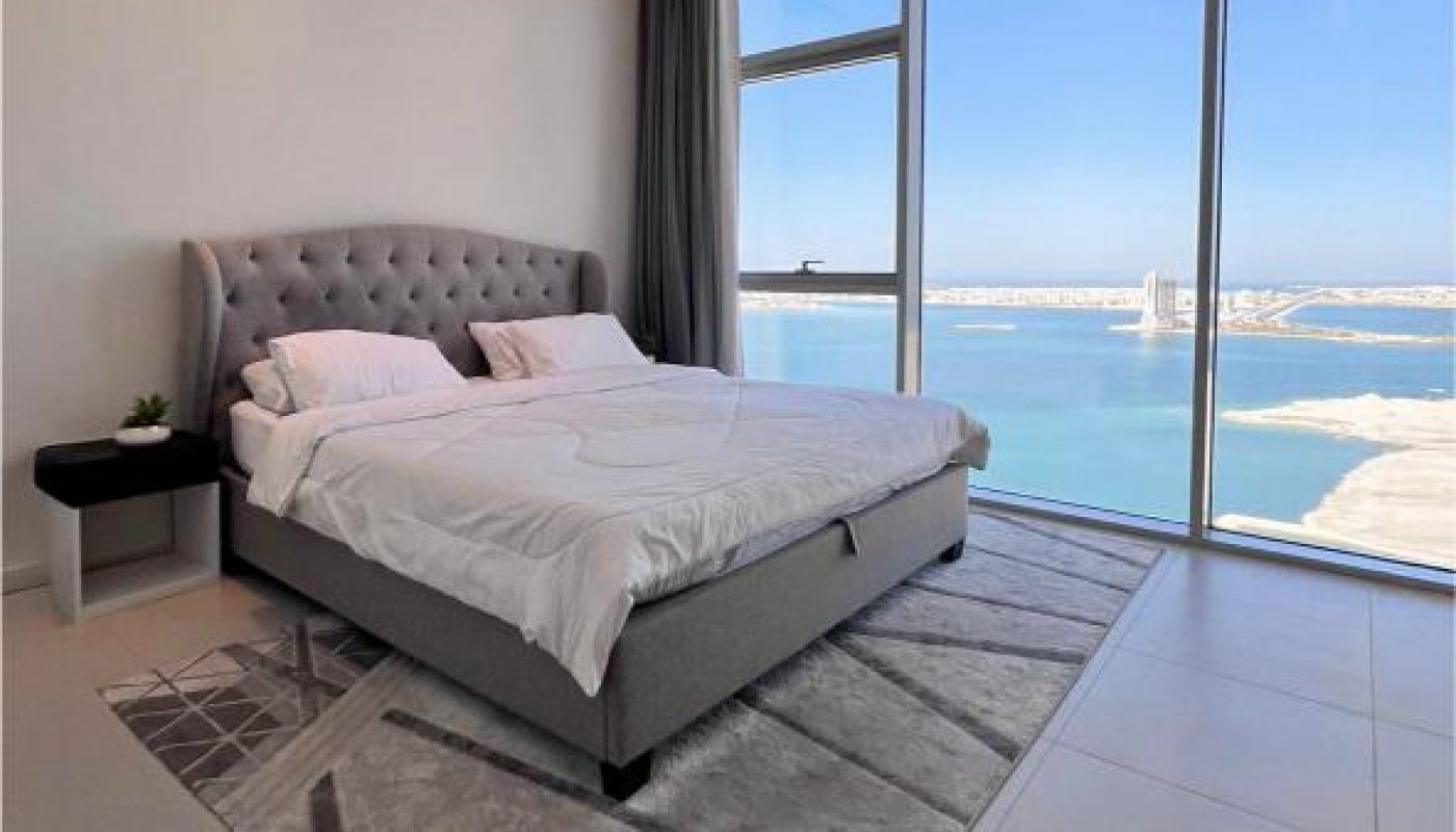 A modern bedroom with a large bed, gray headboard, nightstand, and a floor-to-ceiling window offering a view of the sea and a coastal cityscape.