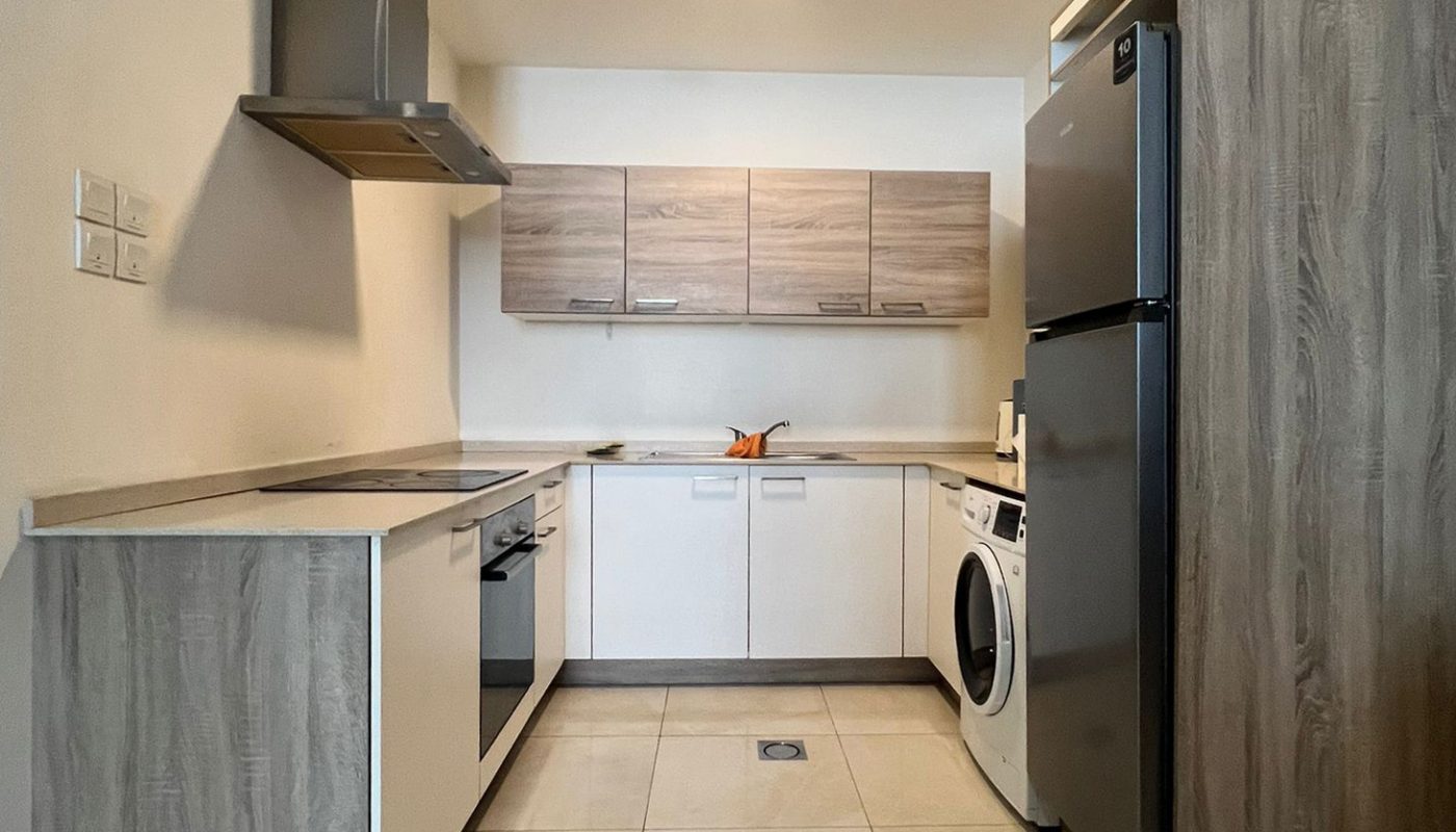 Modern kitchen with light wood cabinets, a stainless steel fridge, washing machine, oven, cooktop, and overhead vent under ceiling lights on a tiled floor.