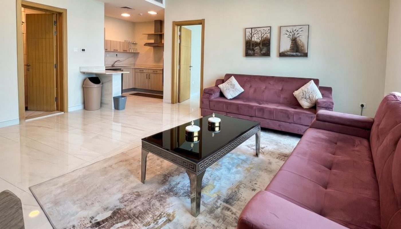 A modern living room with two mauve sofas, a glass coffee table, a rug, wall art, and an open view of the kitchen area under bright ceiling lights.