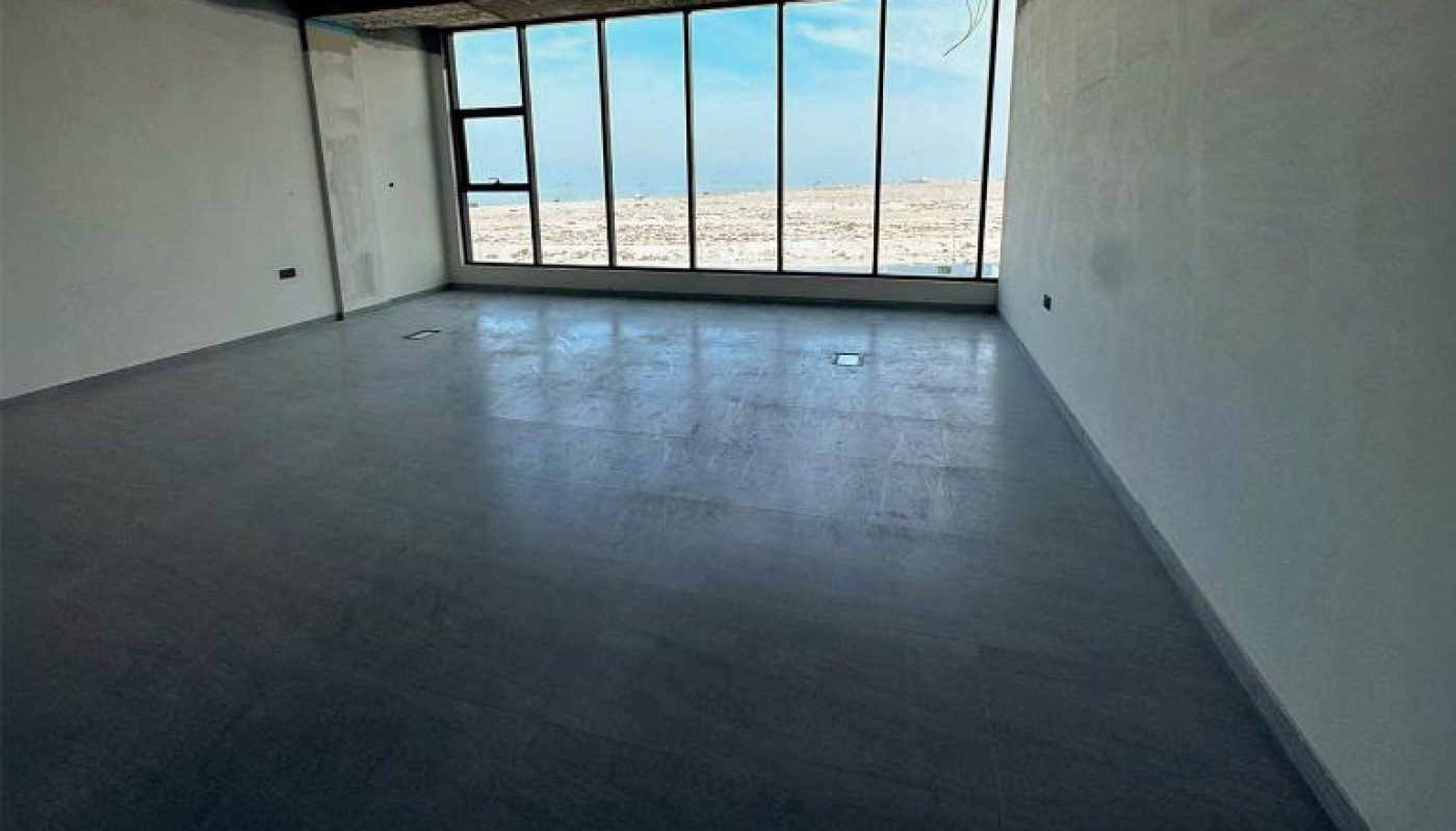 Empty room with dark ceiling, white walls, gray floor, and large floor-to-ceiling windows overlooking a barren landscape under a blue sky.