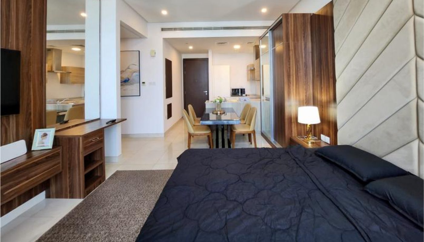 Modern studio apartment with a neatly made bed in the foreground, a dining table and chairs in the center, and a kitchenette and entrance visible in the background.