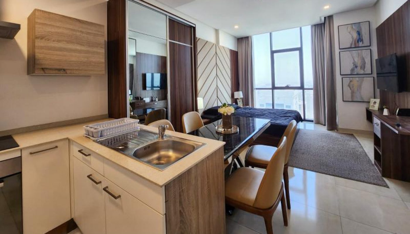 Modern open-plan kitchen and living area with light wood finishes, dining table with chairs, large windows, and a wall-mounted TV.