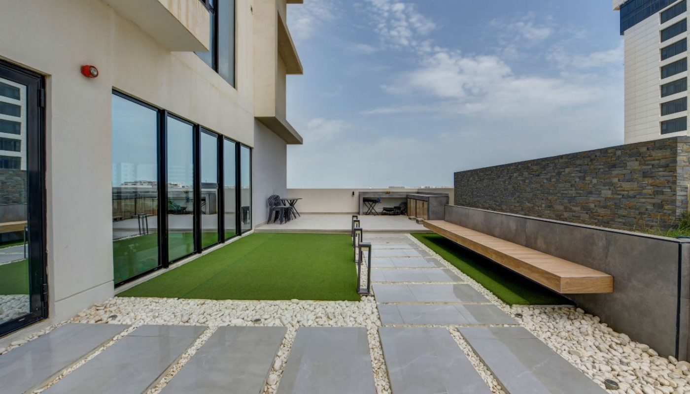 Outdoor terrace with artificial grass, stone pathway, long wooden bench, and glass doors; modern buildings visible in the background under a partly cloudy sky.