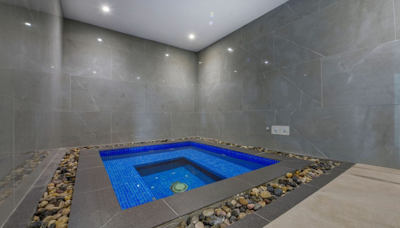 Indoor plunge pool with blue tile surrounded by gray stone walls and a border of river rocks, lit by recessed ceiling lights.