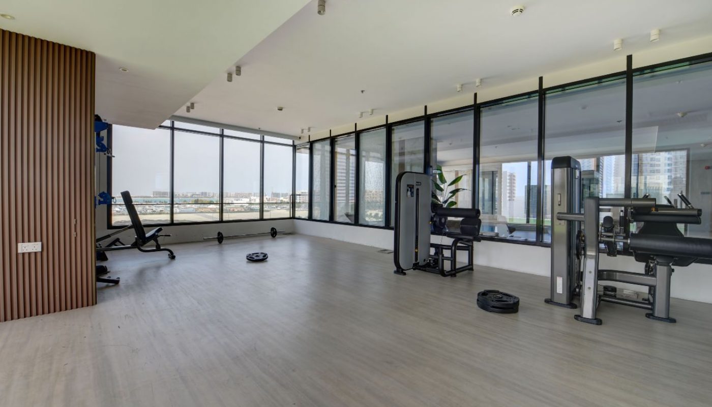 Spacious modern gym with large windows, various weight machines, benches, and free weights on a light-colored floor.