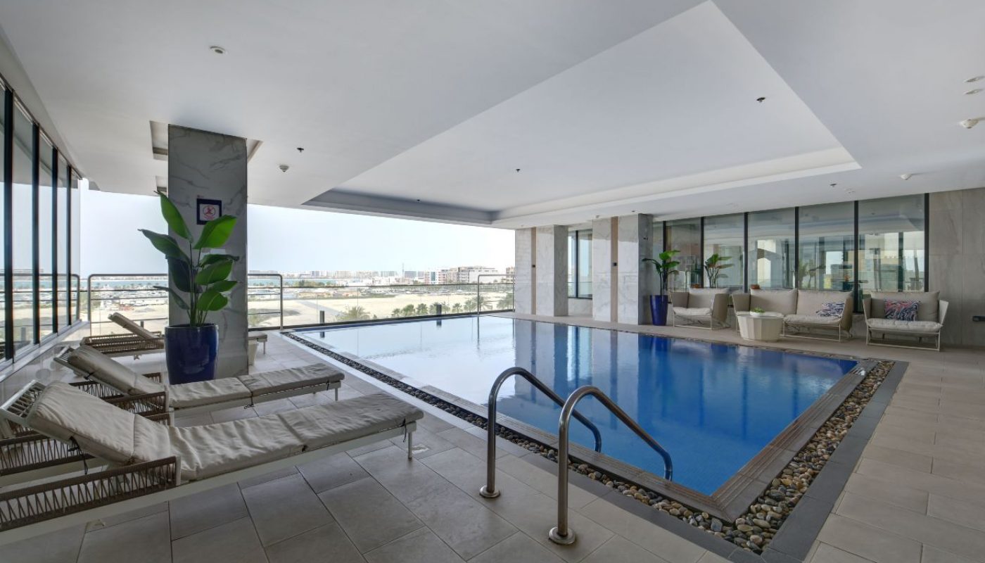 Indoor swimming pool with metal handrails, lounge chairs, potted plants, and large windows offering a city view.