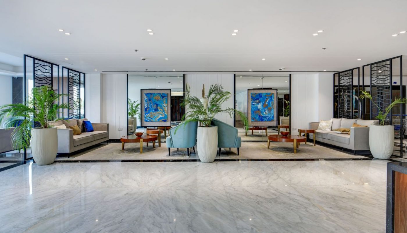 Modern hotel lobby with marble floors, two seating areas with armchairs and sofas, large potted plants, and abstract art on white walls.