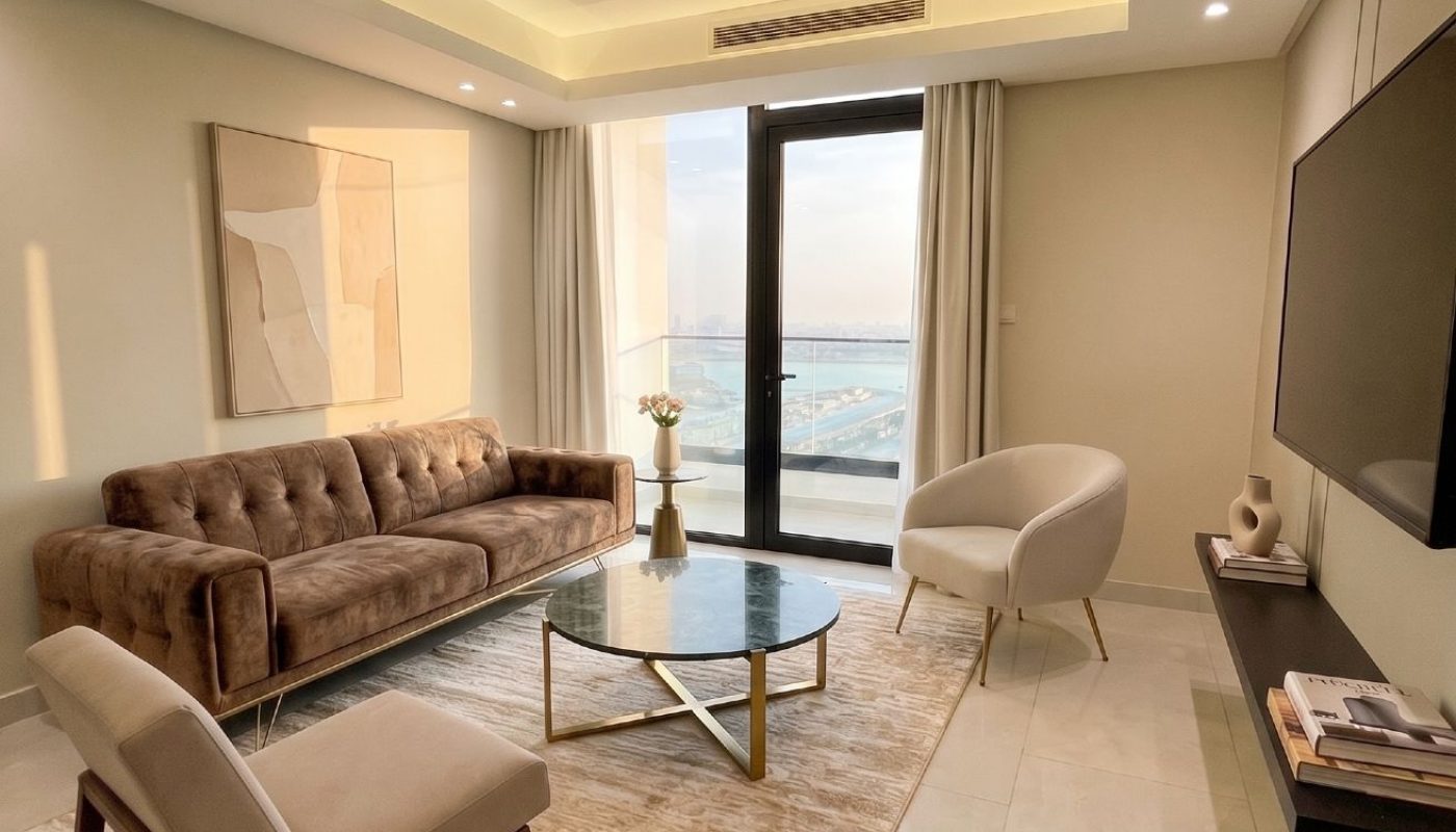 Modern living room with a brown sofa, beige chairs, glass coffee table, abstract wall art, and large window overlooking a waterfront view.