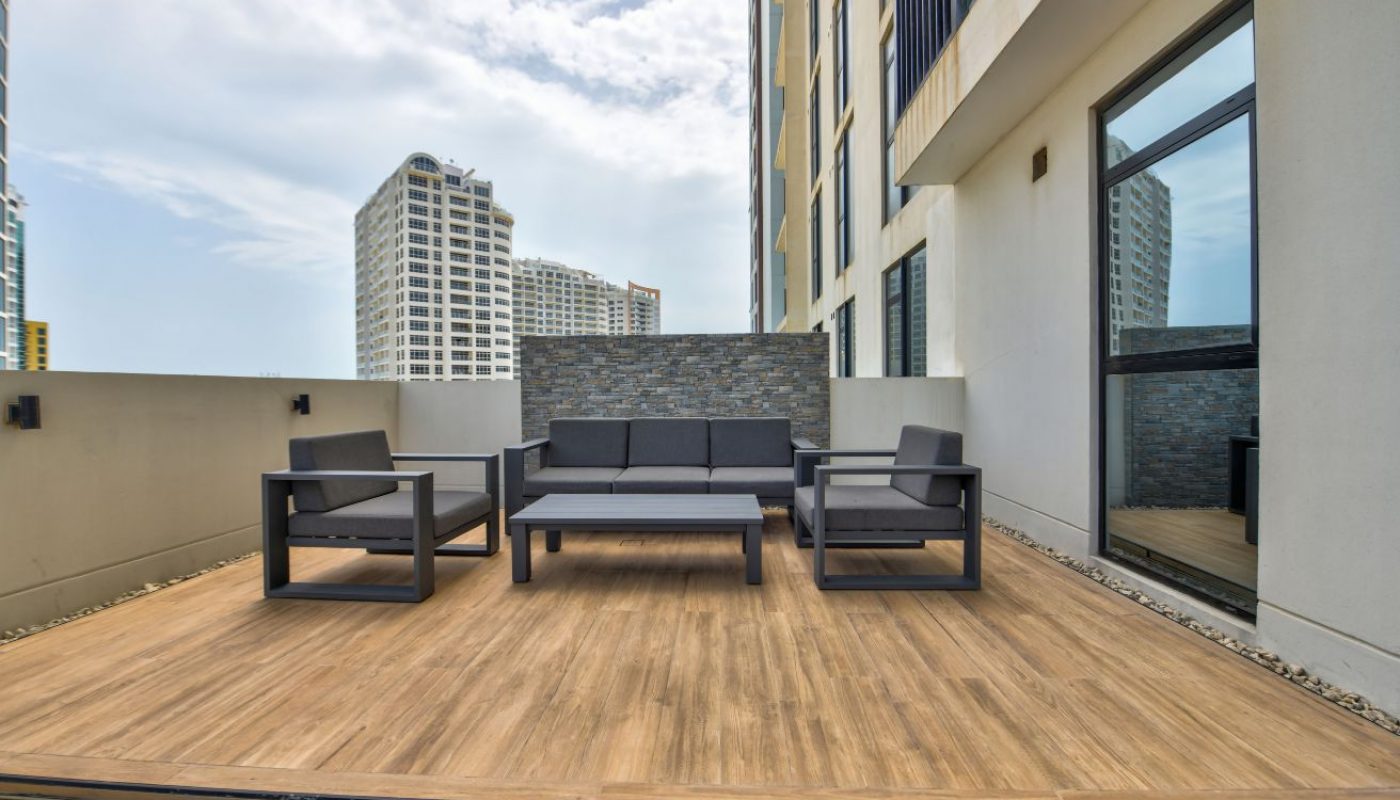 Outdoor patio with wooden deck, gray modern furniture set, and high-rise buildings visible in the background under a partly cloudy sky.