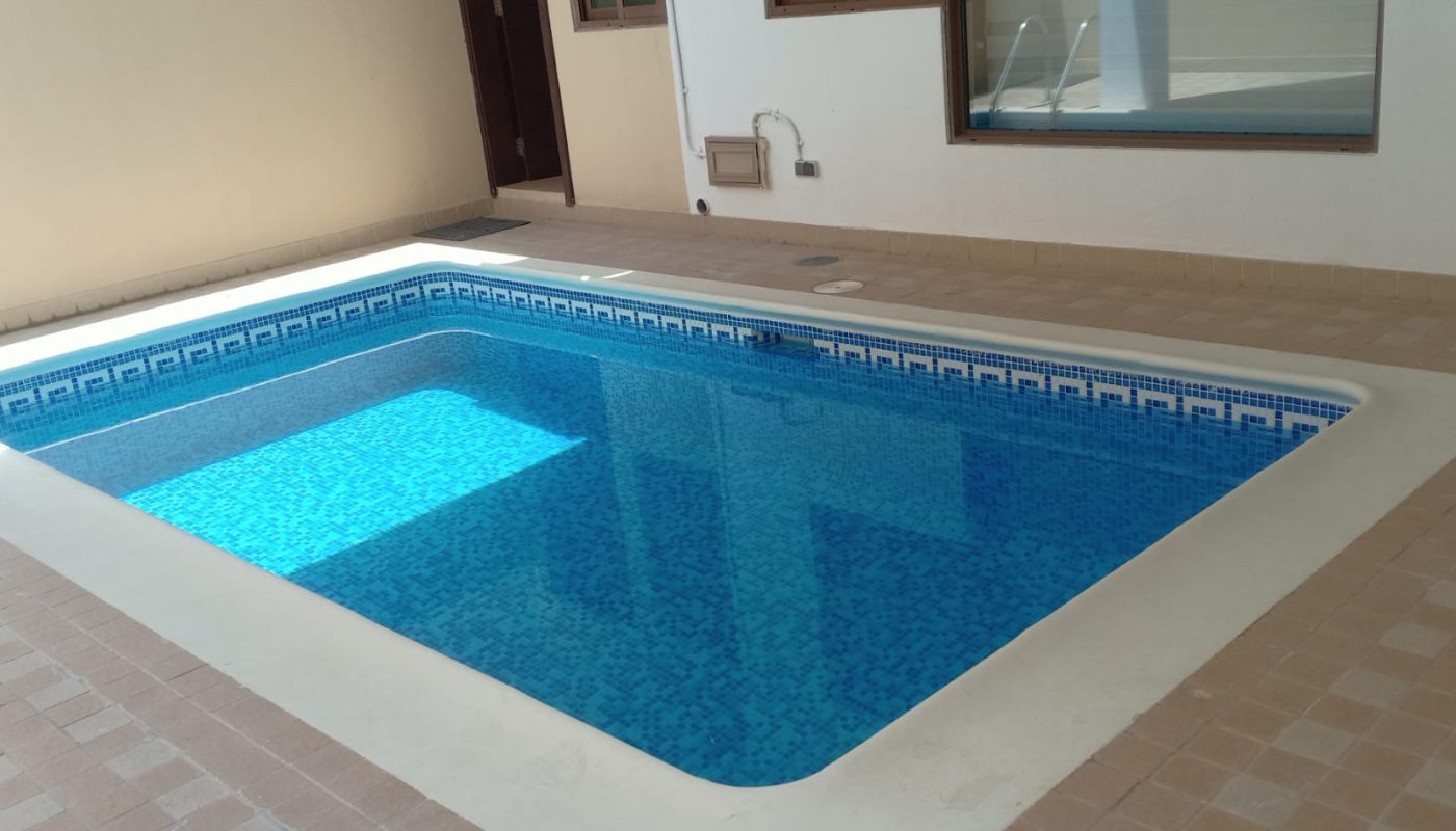 Rectangular indoor swimming pool with clear blue water, surrounded by beige tiles, next to white walls and windows.