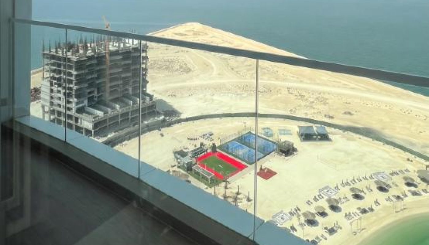 View from a balcony overlooking a construction site, a sports court, a sandy area, and the sea under a clear sky.