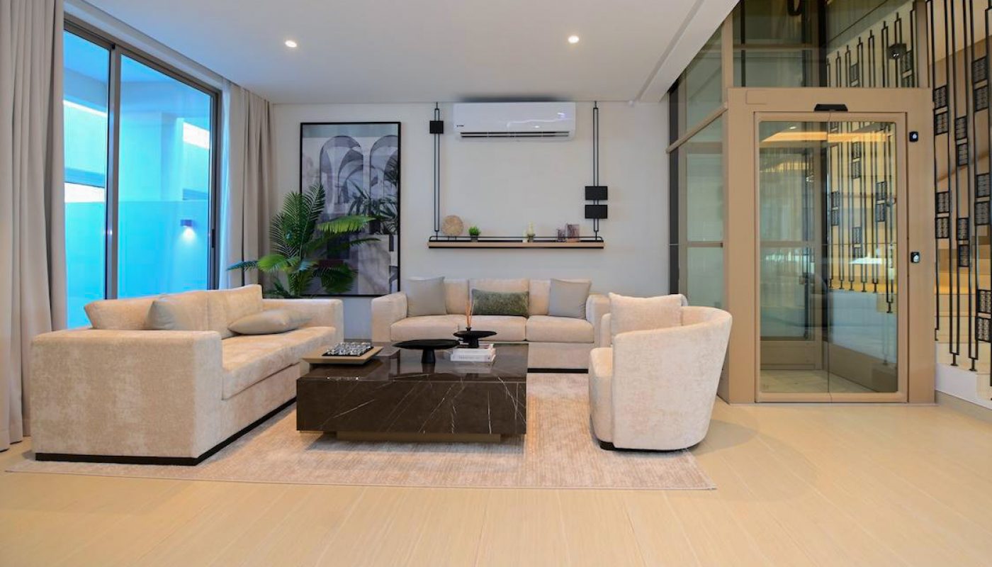 Modern living room with beige sofas, a central coffee table, large windows with curtains, indoor plants, wall shelves, and a glass elevator.