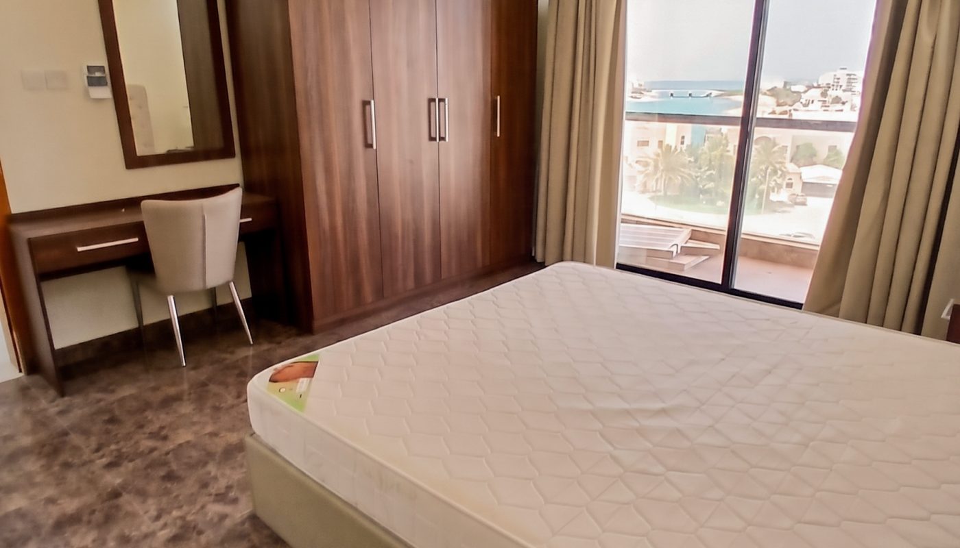 A bedroom with a double bed, wooden wardrobe, desk with chair, large window with curtains, and a view of buildings outside.