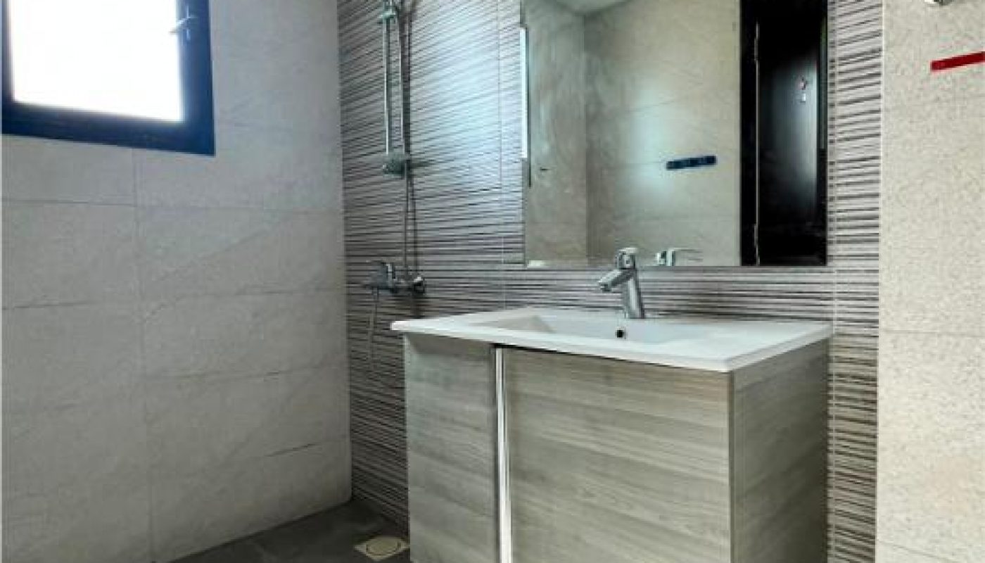 Modern bathroom with a wall-mounted sink cabinet, large mirror, shower area, and a window on the left wall. A pink bucket is visible under the sink.